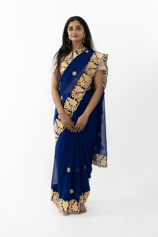 The image shows a person elegantly draped in a navy blue georgette saree. The saree features intricate gold patterns that add a luxurious touch. The fabric is soft and fluid, enhancing the wearer’s natural grace. This saree is perfect for special occasions, exuding sophistication and elegance.