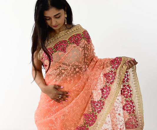  The image shows a person elegantly draped in a ready-to-wear peach net saree in Australia. The saree features pink floral embroidery and detailed golden borders, showcasing meticulous craftsmanship. The soft and lightweight net fabric provides a comfortable and elegant flow. This saree is perfect for special occasions, exuding charm and sophistication.