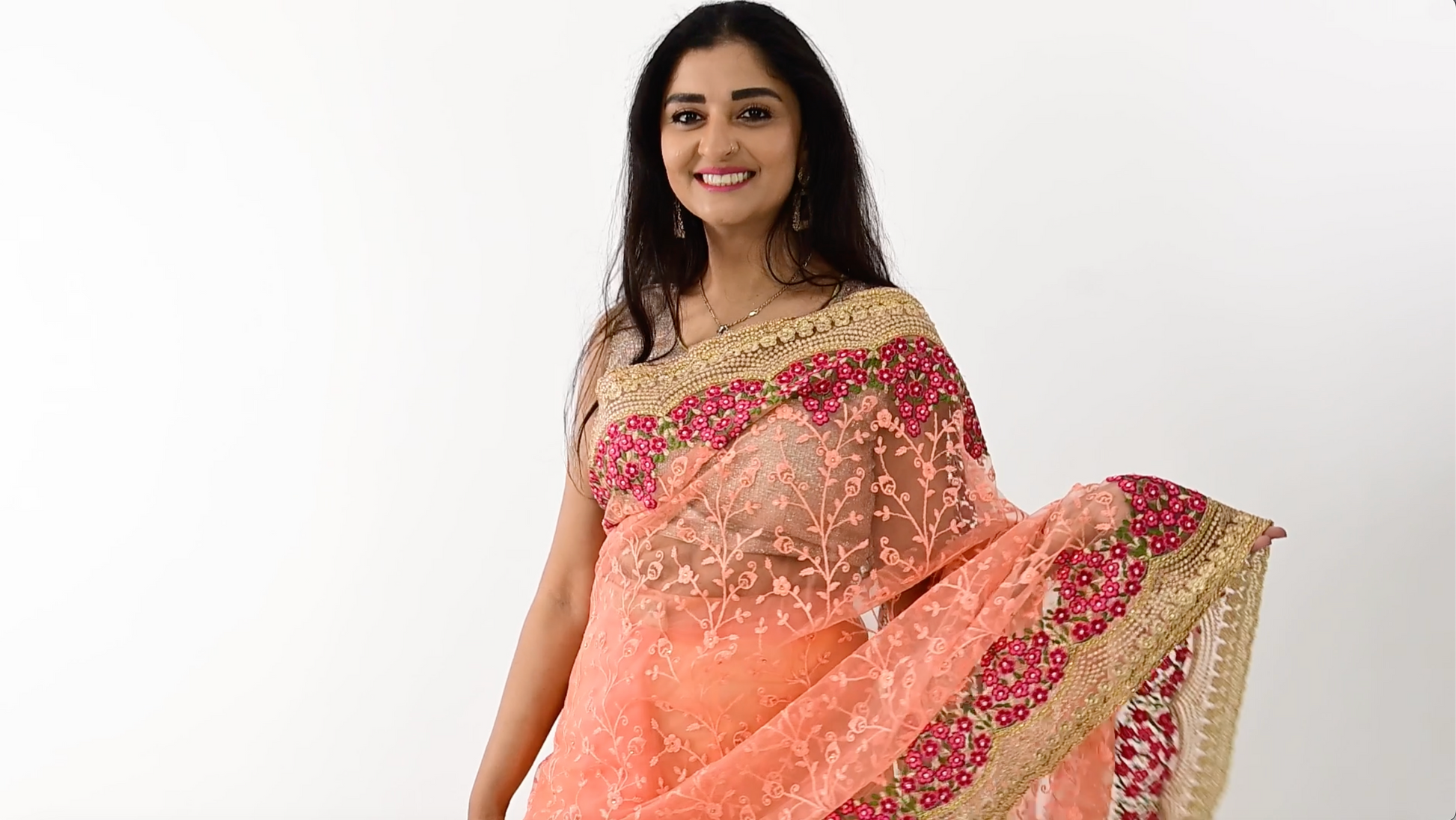 The image shows a person elegantly draped in a ready-to-wear peach net saree in Australia. The saree features pink floral embroidery and detailed golden borders, showcasing meticulous craftsmanship. The soft and lightweight net fabric provides a comfortable and elegant flow. This saree is perfect for special occasions, exuding charm and sophistication.
